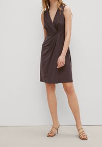 Ärmeloses, dunkelbraunes Kleid mit tiefem V-Ausschnitt, gedrehtem Vorderdesign und knieumspielender Länge aus glattem Stoff. Kombiniert mit nude-farbenen Riemchen-Absätzen.