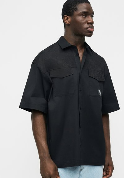 Mladý muž, který má na sobě volnou černou krátkou košili s knoflíky a dvěma kapsami vpředu, a světlé modré džíny, dívá se doprava.