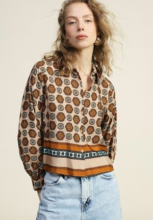 Giovane donna con capelli mossi che indossa orecchini a cerchio dorati, una camicetta a bottoni fantasia marrone e blu e jeans a vita alta azzurri.