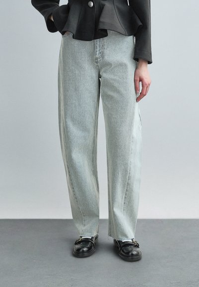 Persona vistiendo jeans sueltos de color gris claro, chaqueta negra abotonada y zapatos de cuero negros, de pie sobre un piso gris contra un fondo gris liso.