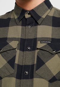 Camisa de franela a cuadros verdes y negros que presenta un cuello con botones, dos bolsillos en el pecho y botones a presión plateados con la etiqueta "CALVIN KLEIN JEANS".