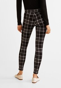 Osoba ubrana w dopasowane, czarno-białe spodnie w kratkę, czarną bluzkę z długim rękawem oraz jasnobeżowe płaskie buty, stoi tyłem do widza.