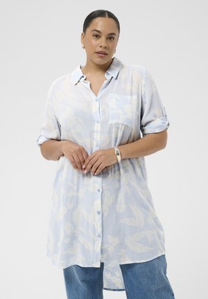 Kvinde med mørkt hår trukket tilbage iført lyseblå blomstret knap-op-skjorte med opsmøgede ærmer og blå jeans, stående mod hvid baggrund.