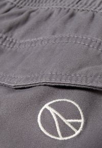 Gros plan sur un tissu gris avec des coutures visibles et un symbole géométrique circulaire blanc brodé en bas.
