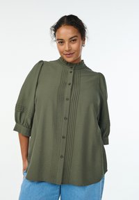 Olivgrön skjorta med hög krage, knappfront och plisserade detaljer. Har puffärmar med subtila randiga accenter. Mjuk, texturerad tyg.