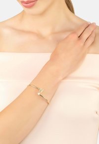 Bracciale rigido color oro con una "N" stilizzata e un piccolo cristallo come accento, superficie liscia e design minimalista.