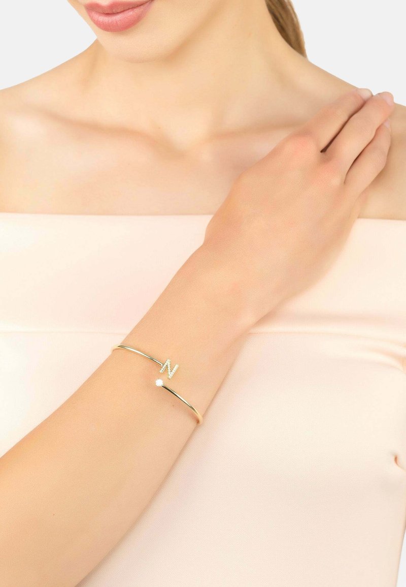 Bracciale rigido color oro con una "N" stilizzata e un piccolo cristallo come accento, superficie liscia e design minimalista.