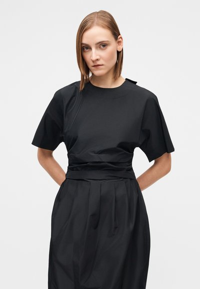 Young woman with straight shoulder-length hair wears a black pleated dress with short sleeves and stands with hands behind her back.
