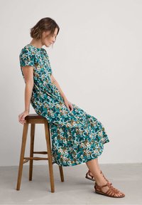 Floral midi dress featuring a teal background, white and brown flower patterns, short sleeves, and a tiered hem, paired with brown strappy sandals.