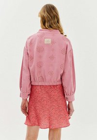 Różowa bawełniana kurtka z kwiatowymi wycięciami i elastycznym pasem, zestawiona z różową spódnicą w kwiatowy wzór. Metka wyświetla nazwę marki "NAOKO."