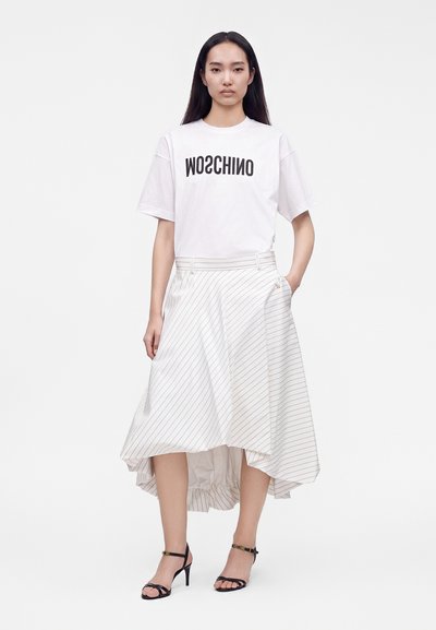 Γυναίκα στέκεται φορώντας ένα λευκό oversized T-shirt με μαύρο κείμενο και μια ασύμμετρη λευκή midi φούστα με ρίγες και μαύρα σανδάλια με τακούνι.
