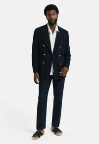 Navy pinstripe double-breasted blazer met gouden knopen, gecombineerd met een licht overhemd, donkere broek en sandalen met een strikaccent.