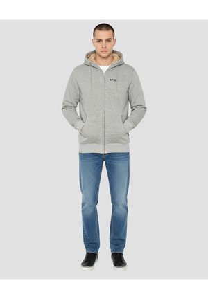 Jasnoszara bluza z kapturem na zamek błyskawiczny o fakturowanym materiale i beżowym kapturze wyściełanym polarem, zestawiona z niebieskimi dżinsami i czarnymi tenisówkami.