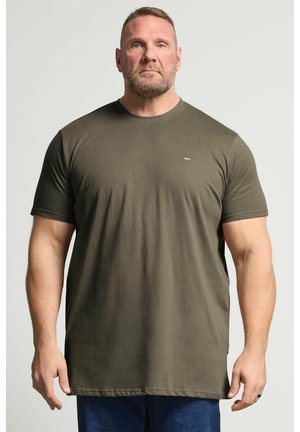 Muskeløs mand med kort hår og skæg iført en olivengrøn t-shirt og blå bukser, stående mod en ensfarvet lys baggrund.