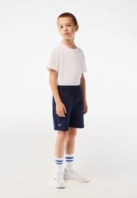 T-shirt bianca in cotone e pantaloni corti blu navy con fascia elastica in vita. Sneakers bianche con dettagli verdi e calze a righe. Dettaglio del logo sui pantaloni.