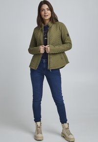 Veste matelassée vert olive avec col montant, poches latérales zippées et motif en chevrons ; associée à un jean foncé et des bottes beiges.