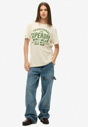 Kremowa koszulka z krótkim rękawem z zielonym nadrukiem. Połączona z luźnymi niebieskimi dżinsami i czarnymi sznurowanymi butami.