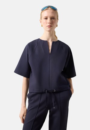 Kobieta nosząca granatowy top z krótkim rękawem z rozcięciem przy dekolcie oraz pasujące spodnie ze ściągaczem w talii, okulary przeciwsłoneczne na głowie i złote kolczyki.