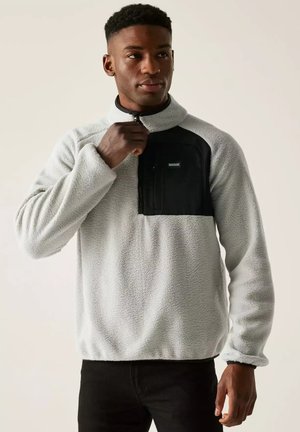 Joven con jersey polar gris claro con bolsillo negro en el pecho, ajustando la cremallera, de pie frente a un fondo liso.