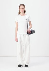 Biele tričko s logom, voľné biele nohavice a čierna crossbody taška. Model nosí čierne slip-on topánky s bielymi podrážkami. Jednoduché pozadie.