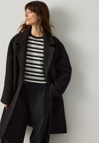 Cappotto nero testurizzato con grandi revers, indossato sopra una maglia a righe bianche e nere. L'outfit è completato da pantaloni neri larghi.