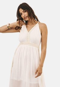 Vestido maxi blanco con cuello halter, que presenta detalles plisados y una cinturilla trenzada. La tela ligera cae hasta los tobillos.