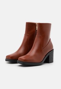 Bottes courtes en cuir marron avec un bout carré, une fermeture éclair latérale et un talon noir épais. Texture lisse avec un design minimaliste.