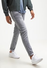 Vaqueros ajustados de color gris claro con un sutil desgaste, dobladillos enrollados y textura suave. Combinados con zapatillas deportivas blancas y una chaqueta bomber oscura.