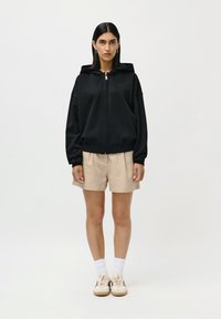 Woman with straight dark hair wearing black zip-up hoodie, beige shorts, white socks, and beige sneakers standing against plain white background.