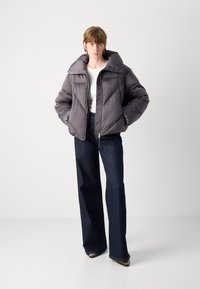 Grå pufferjakke med høy krage og glidelås; quiltet mønster; brukt over en hvit topp og parret med mørke vide jeans.
