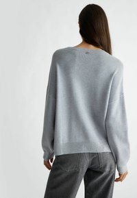 Světle šedý oversized svetr s padlými rameny, žebrovaným lemem a jemnými metalickými detaily na manžetách. Nošený s tmavě šedými džíny.