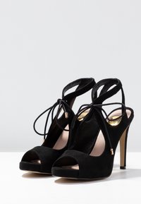 Czarne zamszowe sandały na wysokim obcasie z odkrytymi palcami, zapięcia w kostce związane w kokardki oraz złote akcenty na białej powierzchni.