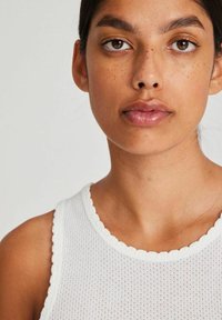Top in tessuto bianco con scollatura a onde. Primo piano del volto di una persona, che mette in risalto il tono della pelle naturale e le lentiggini.