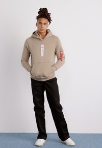 Beige hettegenser med frontlomme og logo, svarte bukser og hvite joggesko. Modellen står mot en ensfarget bakgrunn.