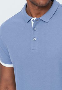 Světle modrá polo tričko s límečkem, zapínáním na tři knoflíky a bílými akcenty na rukávech. Vyrobeno z texturované tkaniny.