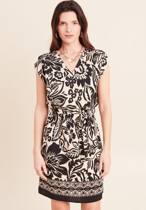 Frau mit langen braunen Haaren trägt ein ärmelloses beiges Kleid mit großem schwarzem Blumenmuster, einen Taillengürtel, goldene Creolen und eine goldene Herz-Halskette.