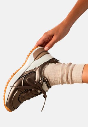 Zapatillas marrones con un acabado brillante, soporte acolchado para el tobillo, entresuela blanca y suela naranja; los cordones atados y un calcetín beige visible.