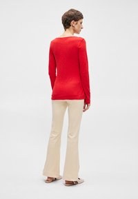 Vörös, hosszú ujjú felső, texturált felülettel, krém színű, bővülő nadrággal és világos szandállal párosítva, hátulról nézve.