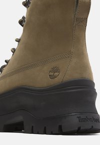 Brown suede hiking boot with a black rubber sole, metal eyelets, and a tree logo. Features a padded collar and textured surface.