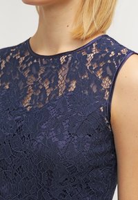 Navy blue lace dress with floral pattern, sheer neckline, and solid fabric underneath. Satin detailing around the armholes.