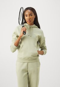 Sweatshirt com capuz de verde claro e calças de fato a condizer, com um bolso frontal e detalhes em logo dourado, segurado com uma raquete de ténis.