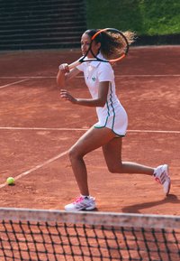 White tennis dress with green accents, textured court surface, player holding a racket, pink and white tennis shoes, yellow tennis ball nearby.