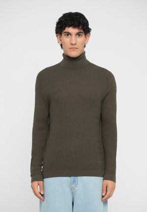 Pulover mulat, cu guler de tip turtle, de culoare verde închis, cu un model texturat și cusături, purtat cu blugi largi, de culoare albastru deschis.