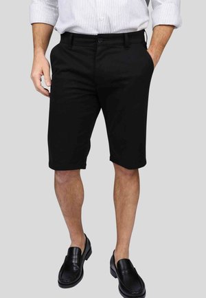 Shorts en coton noirs à coupe droite, longueur genou, avec une fermeture à bouton unique et des poches latérales, portés avec des mocassins noirs.