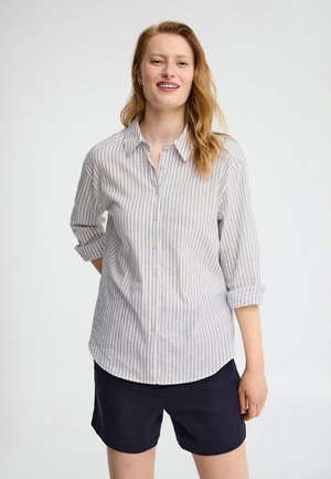 Lächelnde Frau mit langen roten Haaren trägt ein weiß-grau gestreiftes Hemd mit Knöpfen und schwarze Shorts, steht vor einem einfarbigen weißen Hintergrund.