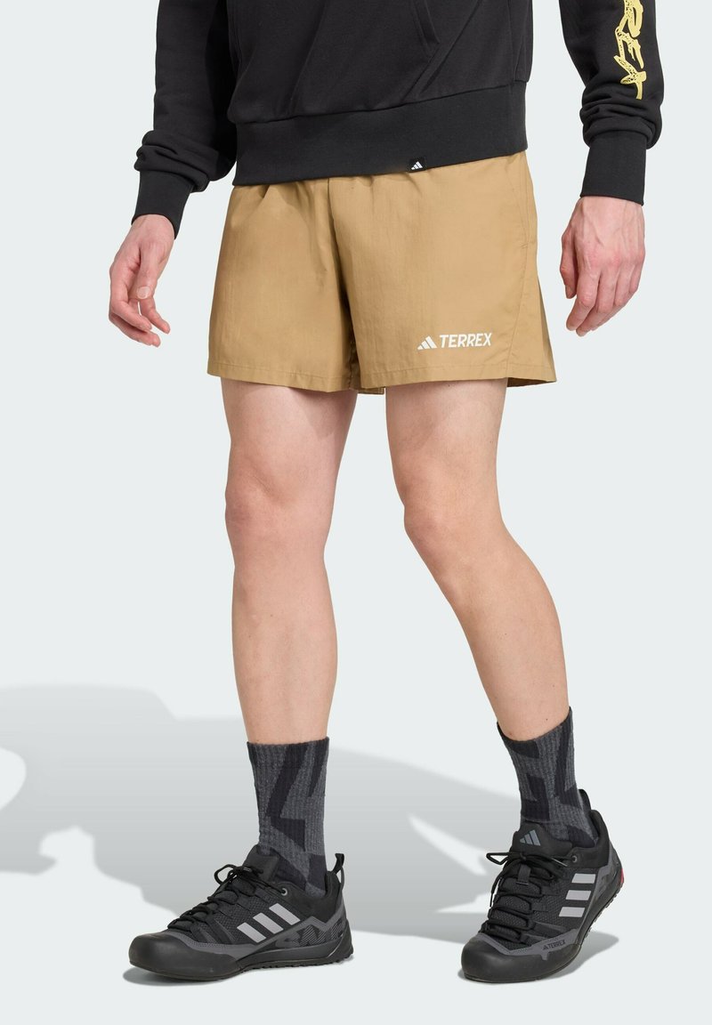 Tanfarbene Sportshorts mit einem elastischen Bund, weißem "TERREX"-Logo, kombiniert mit schwarzen Schuhen und dunkel gemusterten Socken. Glattes, leichtes Material.