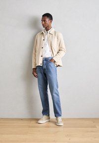 Giacca beige con bottoni neri sopra una camicia bianca, abbinata a jeans blu chiaro e sneakers beige, in piedi su un pavimento di legno.