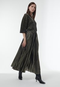 Robe longue plissée en vert foncé avec un éclat, présentant un décolleté en V et des manches trois-quarts. Portée avec des bottes à talons hauts noires.