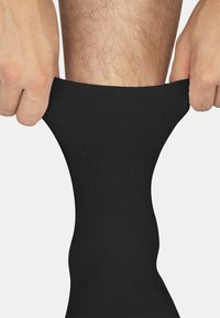 Schwarze gerippte Socke, die hochgezogen wird, während Hände den oberen Teil greifen. Die Socke hat eine glatte Textur und einen engen Sitz.