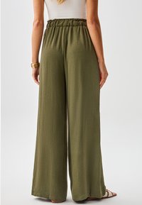 Pantaloni a gamba larga in tessuto verde oliva, con superficie testurizzata e vita elastica. Sandali visibili nella parte inferiore.